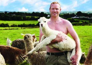 Cute, shirtless redheaded Irish farmer, holding a baby alpaca. Other alpacas surround him.