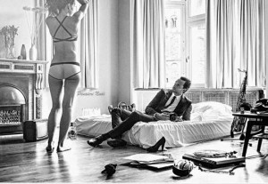 Man in suit reclines on bed watching a beautiful woman in lingerie. A saxaphone is propped by the bed. 